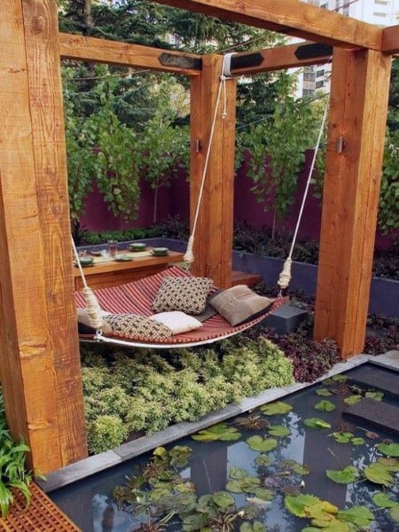 Outdoor hammock bed with cushions, wooden pergola, and a serene pond surrounded by greenery.