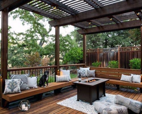 Cozy pergola with wooden seating, fire pit table, string lights, and a lush outdoor backdrop.