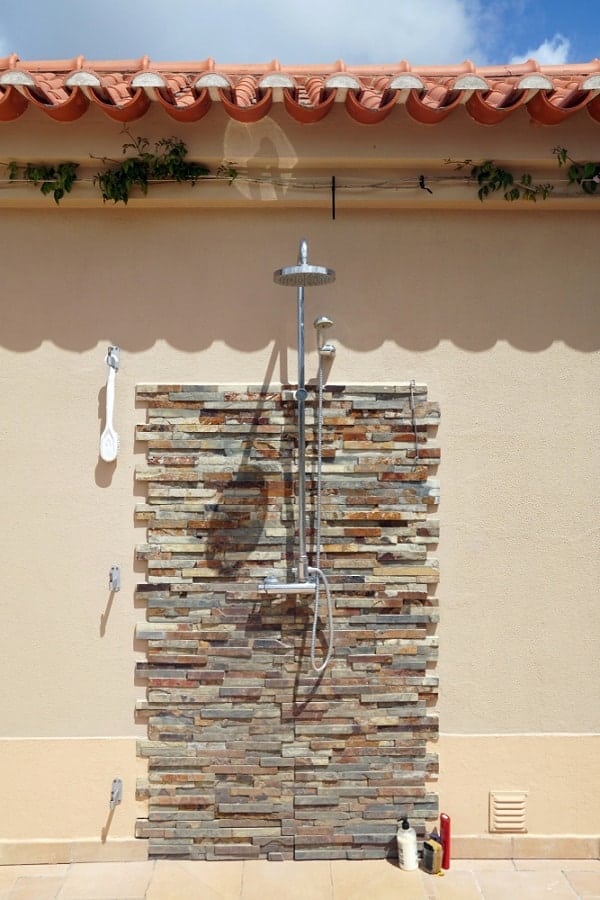 Outdoor shower with a textured stone wall, beige backdrop, and terracotta roof tiles.