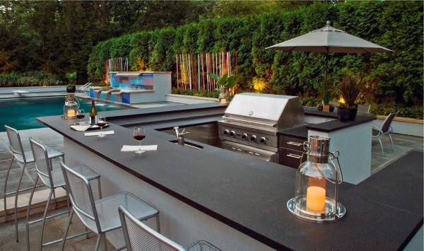 Modern outdoor kitchen with a grill, bar seating, candle lanterns, and an inviting outdoor pool in the background