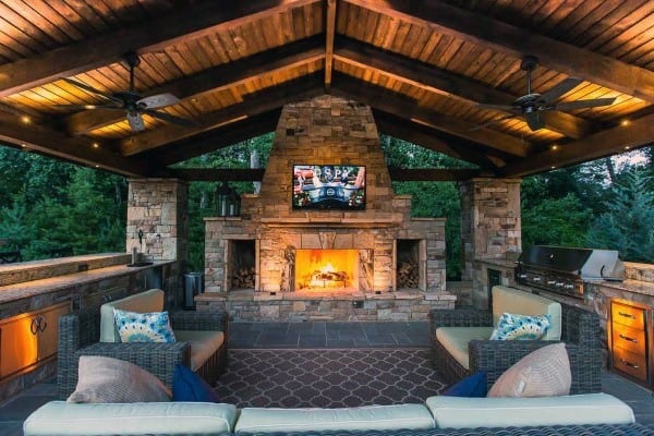 An outdoor patio with a stone fireplace, TV, seating area, wooden ceiling, surrounded by trees and ambient lighting