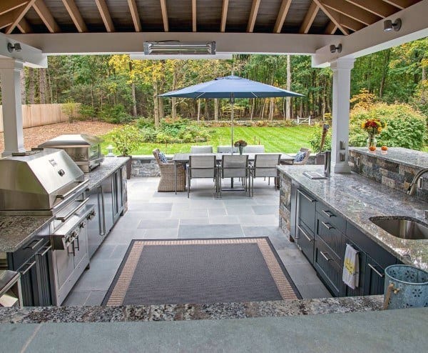 Outdoor kitchen with stainless appliances, granite countertops, and a dining area under an outdoor blue umbrella in a garden