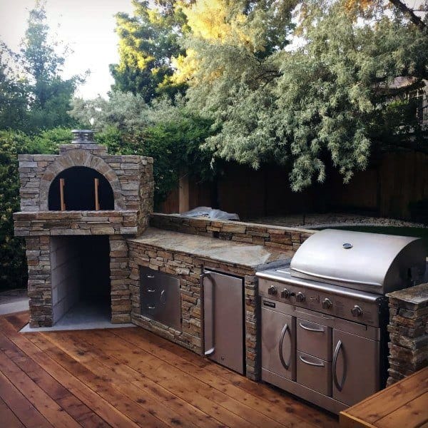 Outdoor kitchen featuring a stone oven, stainless grill, and wooden deck, all nestled in lush greenery