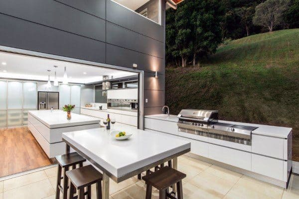 Modern outdoor kitchen with sleek white countertops, stainless steel appliances, and adjacent greenery
