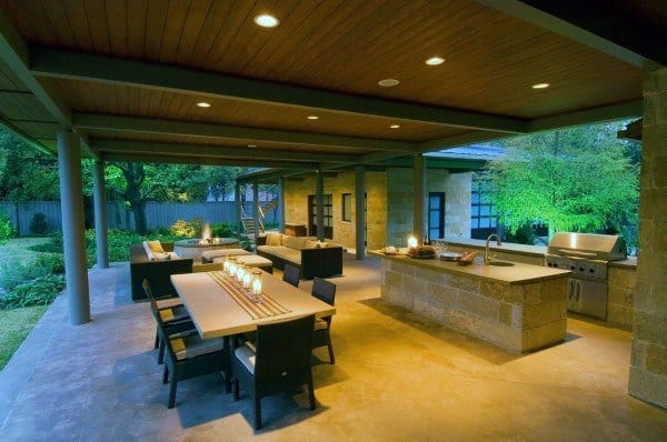 Outdoor covered patio with dining area, sofas, and a kitchen setup, featuring warm lighting and garden views