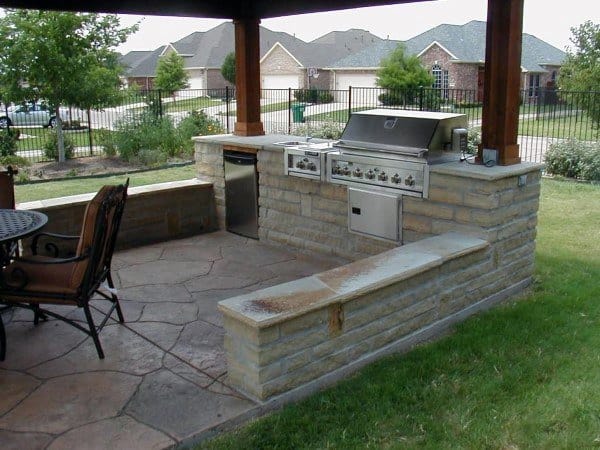 Outdoor kitchen with a stone grill island and dining area under a wooden pergola in an outdoor suburban backyard