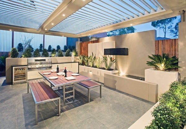 Modern outdoor kitchen with a pergola, grill, seating area, and plants on a tiled outdoor patio