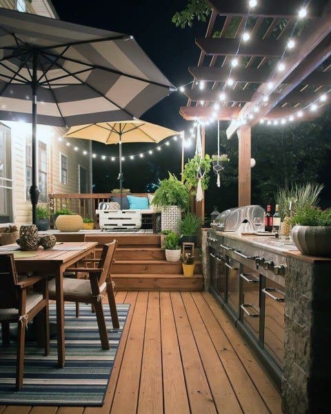 Cozy outdoor patio at night with string lights, umbrellas, dining area, and a grill on a wooden outdoor deck