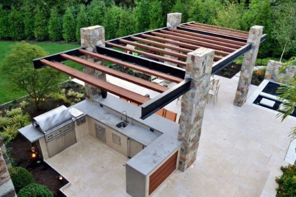 Outdoor kitchen with pergola, stone pillars, stainless steel appliances surrounded by lush outdoor greenery