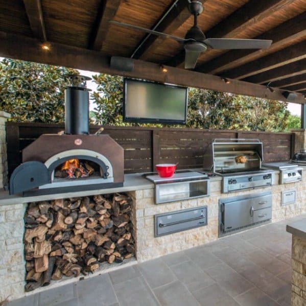 Outdoor kitchen with a built-in grill, wood-fired oven, TV, and stacked firewood under a pergola