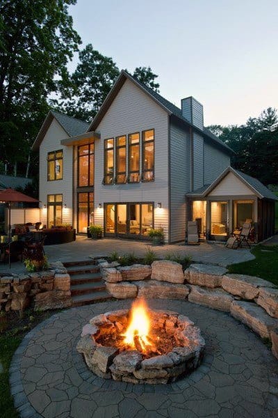 Modern house with large windows, a patio, and a stone fire pit surrounded by lush greenery