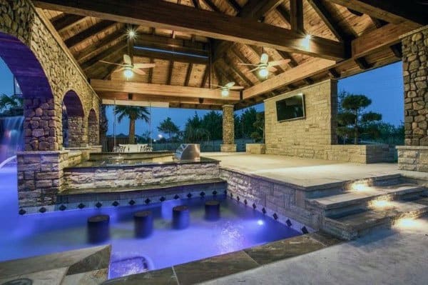 Outdoor stone patio with poolside bar and built-in seating under a wooden pergola.