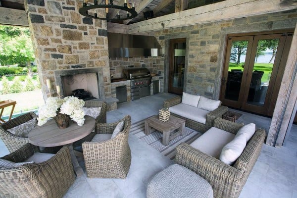Outdoor patio with wicker furniture, a cozy fireplace, grill, and stone walls; lush greenery visible through glass doors