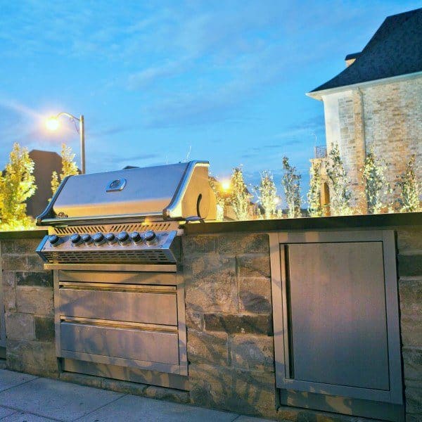 Outdoor stainless steel grill seamlessly integrated into a stone counter at dusk, with lights and house in view
