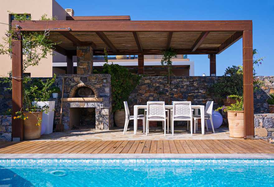 Poolside terrace with a wood pergola, stone pizza oven, and dining table with white chairs.