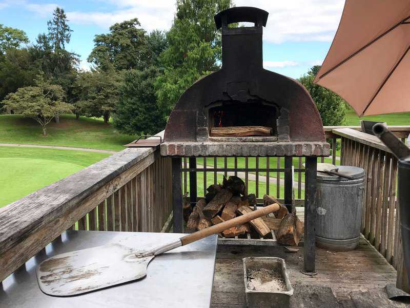 Rustic outdoor wood-fired pizza oven on a balcony with firewood and pizza peel nearby.
