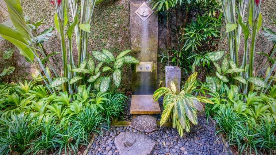 Rustic outdoor shower with a mossy stone wall, pebble flooring, and lush tropical plants.