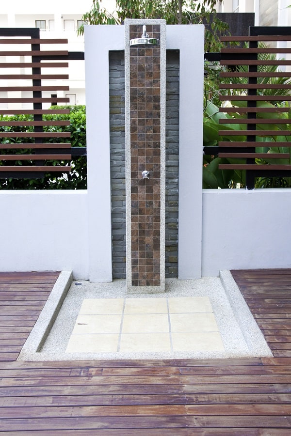 Modern outdoor shower with tiled wall and floor, framed by wooden decking and greenery.