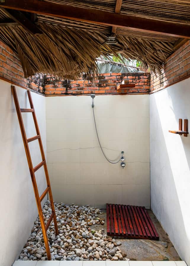 Rustic outdoor shower with pebble flooring, wooden mat, ladder, and thatched roof design.