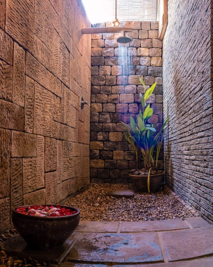 Rustic outdoor shower with stone walls, a potted plant, and a bowl of flower petals.