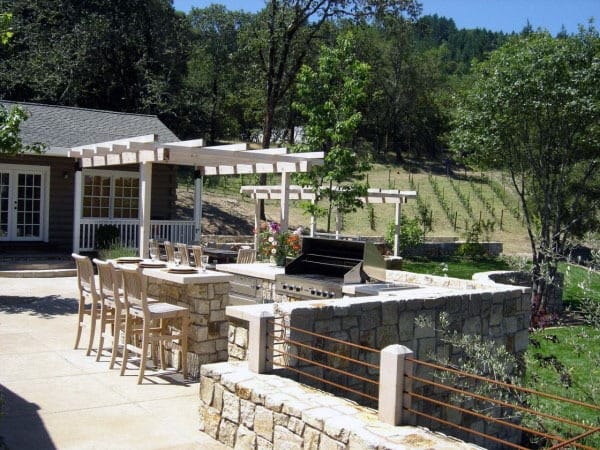 Outdoor stone bar with built-in grill, pergola, and wooden barstools on a spacious patio.