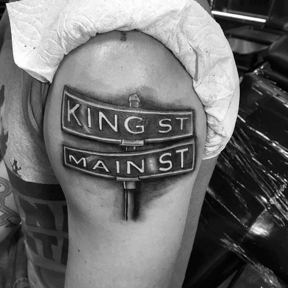 Black and grey tattoo of a realistic street signpost featuring 'King St' and 'Main St,' placed on the upper arm with bold shading.