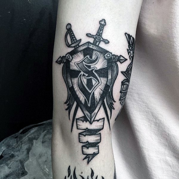 Black ink tattoo on inner forearm of a stylized shield with a bold emblem, crossed swords, and a blank scroll banner beneath it.