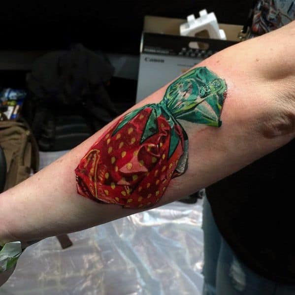 Hyperrealistic strawberry candy wrapper tattoo with intricate shading and highlights, creating a striking 3D effect on the forearm.