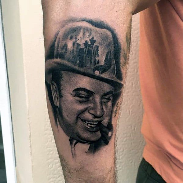 Al Capone tattoo on the forearm, featuring a detailed black-and-white portrait of the gangster smiling with a cigar, with a reflection of a vintage gangster scene in his fedora.