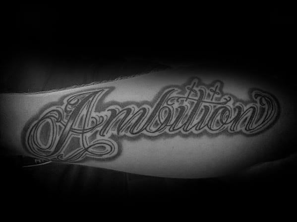 Tattoo of "Ambition" in elegant script graces an arm, contrasted by a shadowy backdrop