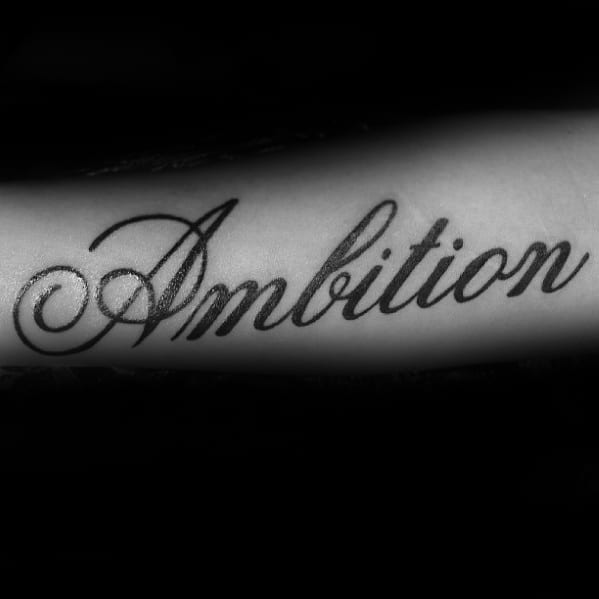 Tattoo scripted "Ambition" elegantly graces an arm, set against a deep, shadowy backdrop
