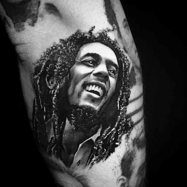 Realistic Bob Marley tattoo with detailed dreadlocks and a bright smile, inked in black and gray on the inner bicep.