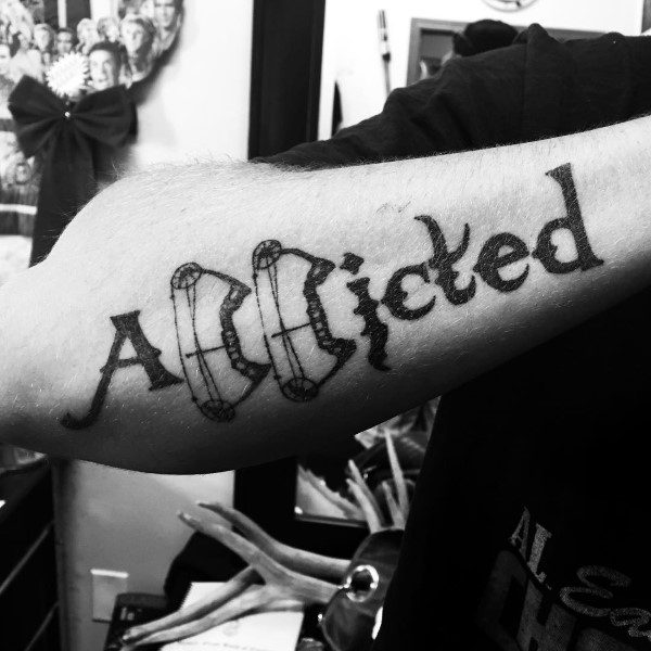 Forearm tattoo of the word Addicted with the A creatively designed using compound bow cams and string elements.
