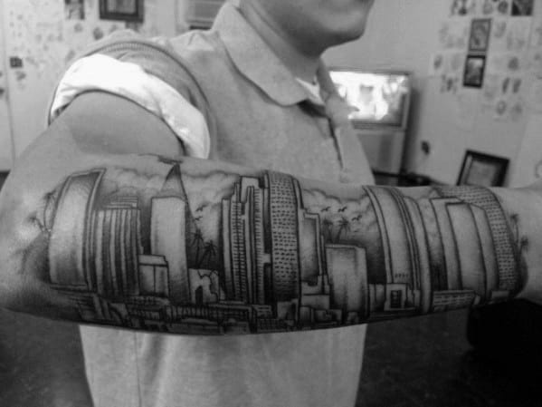 Detailed black and gray forearm tattoo of the Los Angeles skyline with cloud shading and prominent round and rectangular buildings.