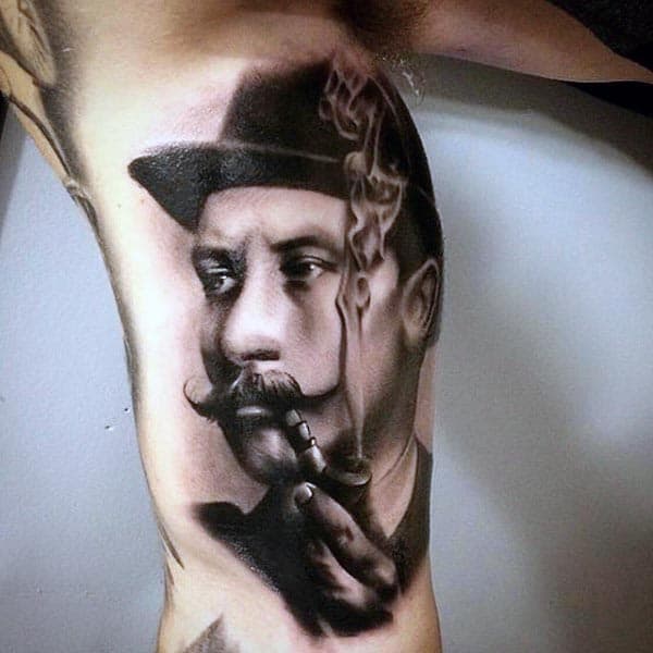 Realistic black and grey tattoo of a distinguished man with a mustache, wearing a hat and smoking a pipe, with detailed shading and smoke effects