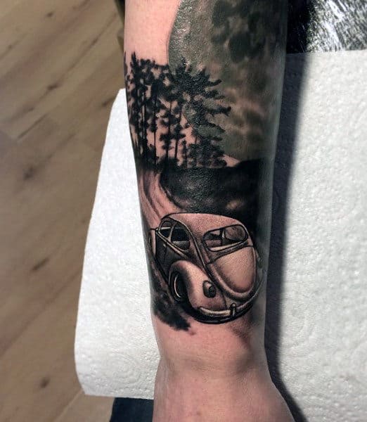 Tattoo of a vintage car on a road surrounded by trees, inked on a forearm