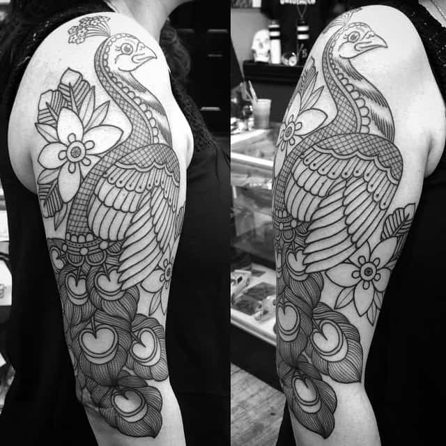 Black and white peacock tattoo with detailed feathers and floral elements, covering the upper arm.