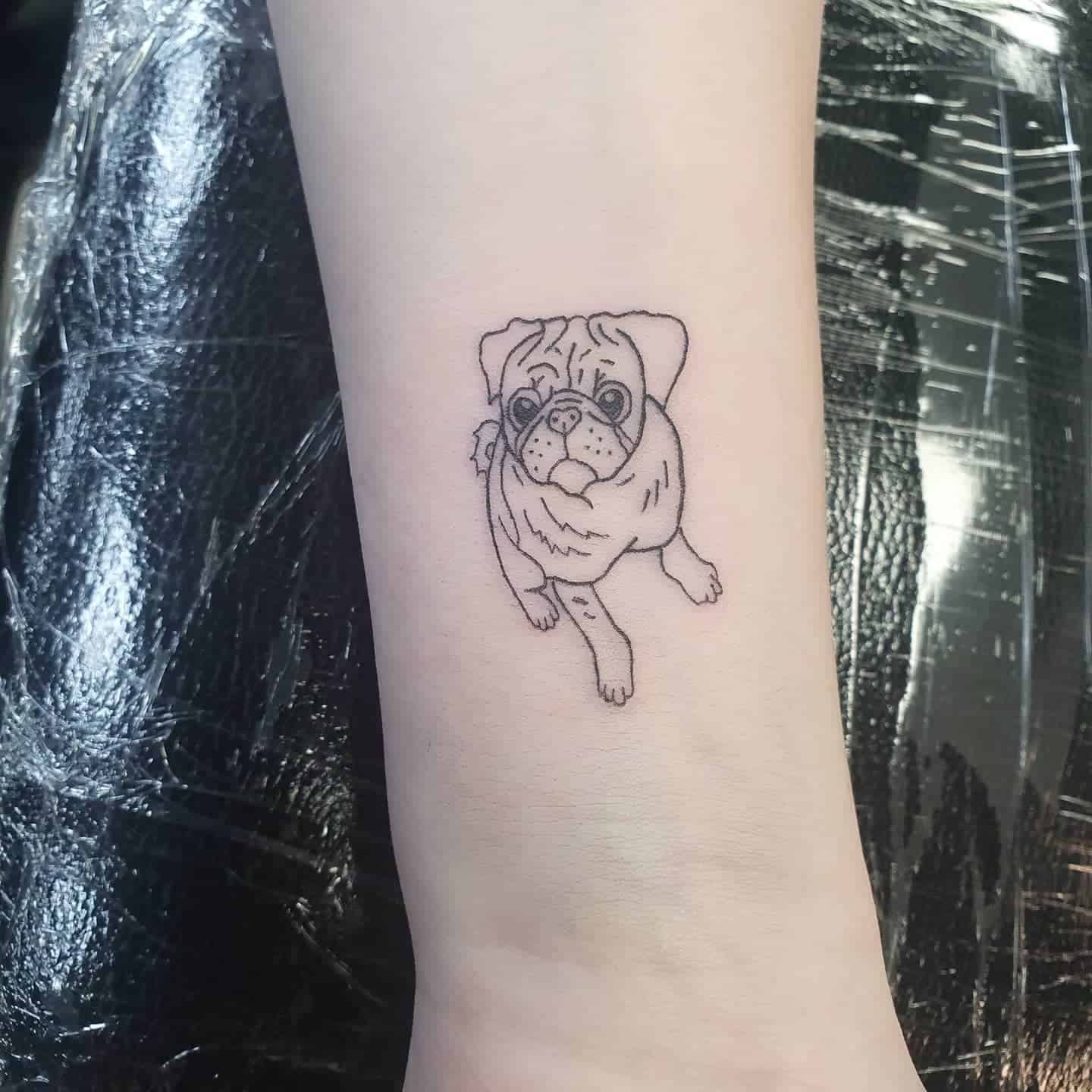 A minimalist line tattoo of a pug looking up with its front paw raised, capturing the dog's curious expression and distinctive features