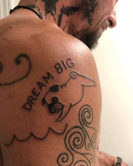 A tattoo featuring the phrase "DREAM BIG" above a playful pug with its ears flapping in the wind, surrounded by flowing lines resembling waves
