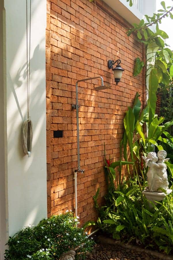 Outdoor shower featuring a rustic red brick wall, tropical greenery, and a decorative statue for an elegant touch.