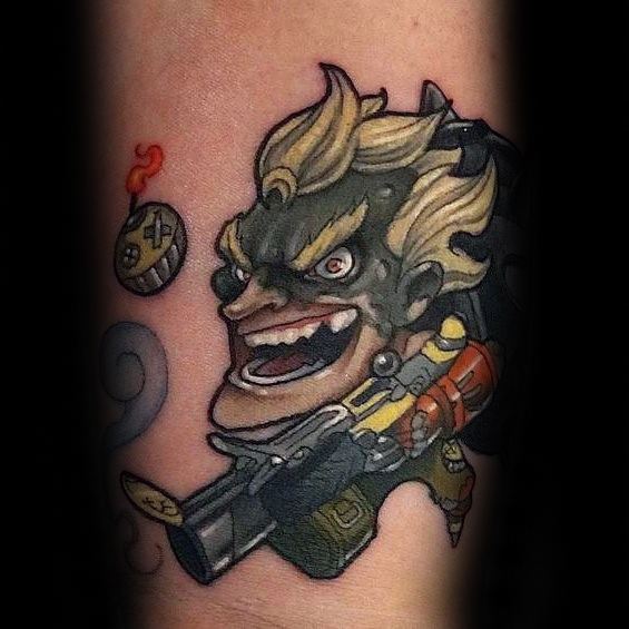Cartoon-style tattoo of Junkrat from Overwatch with his signature grin, grenade launcher, and a flaming bomb on the forearm.
