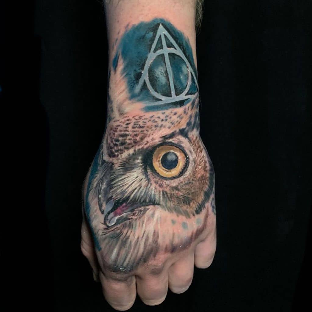 Realistic owl tattoo on the hand with piercing yellow eyes, blue shading, and the Deathly Hallows symbol integrated into the design.