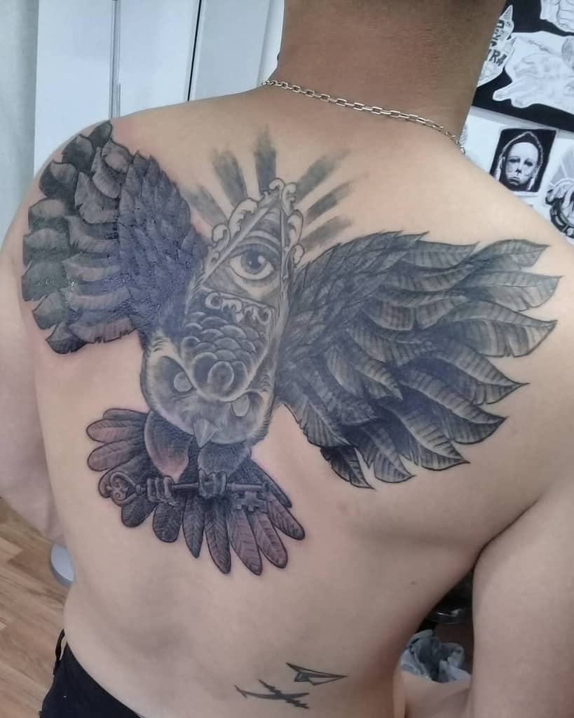 Tattoo of a large owl with an all-seeing eye on its forehead, holding a key in its talons, inked in black and grey shading across the back.