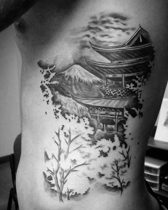 Black and grey side rib tattoo of a Japanese temple with Mount Fuji in the background, surrounded by scattered leaves and branches.