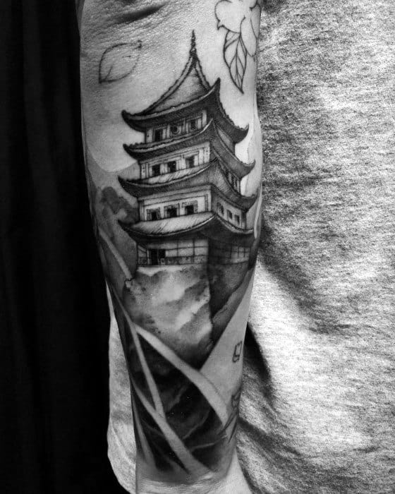 Black and grey forearm tattoo of a tall Japanese pagoda with soft watercolor-style shading, foliage, and falling leaves for a serene look.