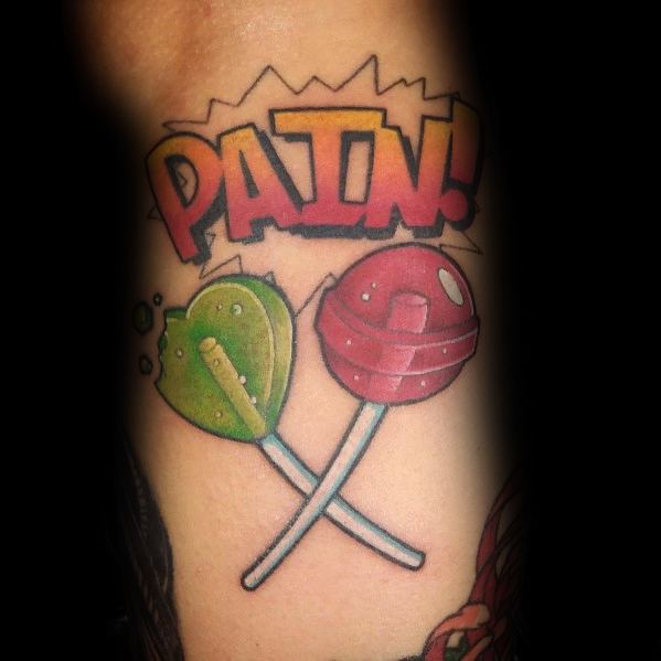 Bold and colorful tattoo featuring crossed lollipops, one heart-shaped and cracked, with "PAIN!" in comic-style lettering above them.