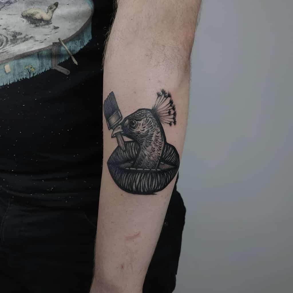Black and gray tattoo of a peacock inside a bowl, with a paintbrush in its beak, located on the forearm.