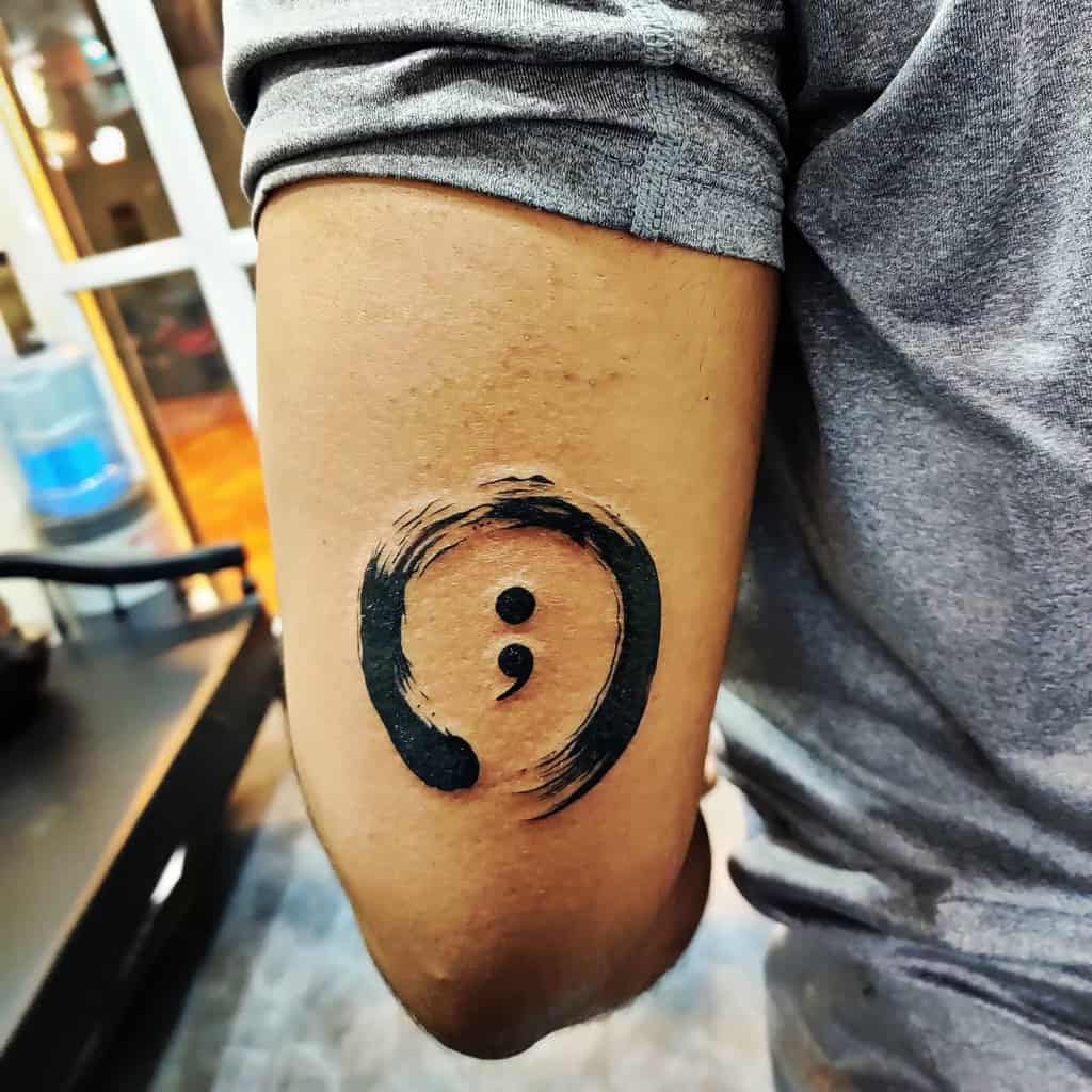 This tattoo features a semicolon enclosed within an artistic circle, symbolizing resilience, continuity, and personal growth. The design incorporates bold, expressive brushstrokes for added impact.
