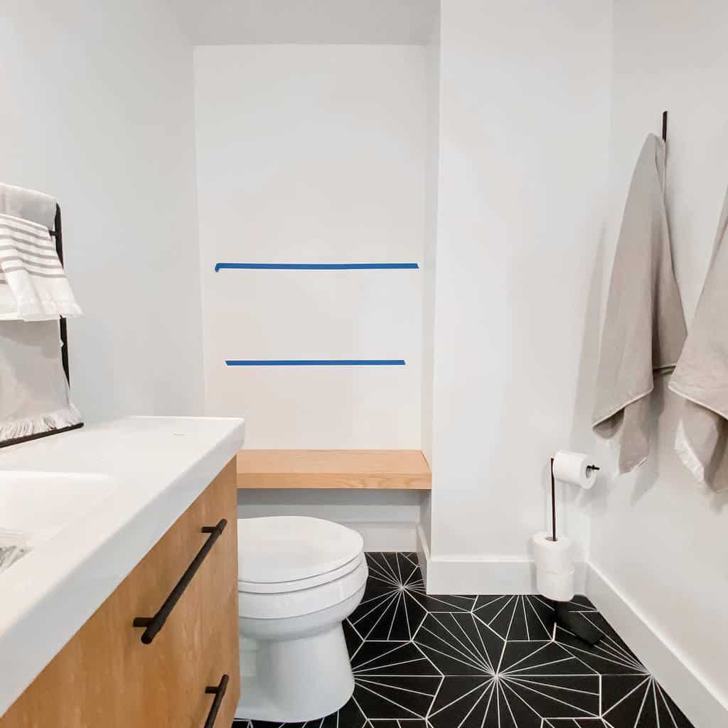 White bathroom with black geometric pattern floor tiles and a minimalist wooden bench.