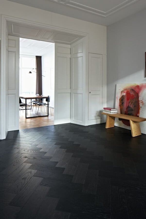 Room with a black herringbone wood floor, white paneled walls, and a wooden bench displaying abstract art.